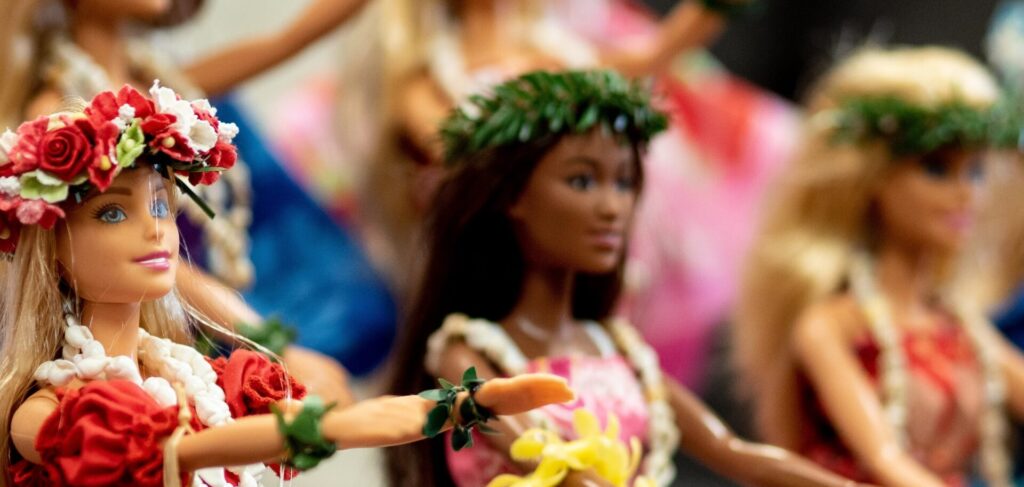 A line of Barbie dolls, some white and some Black, are dressed up as Hawaiian dancers in colourful dresses and flower leis